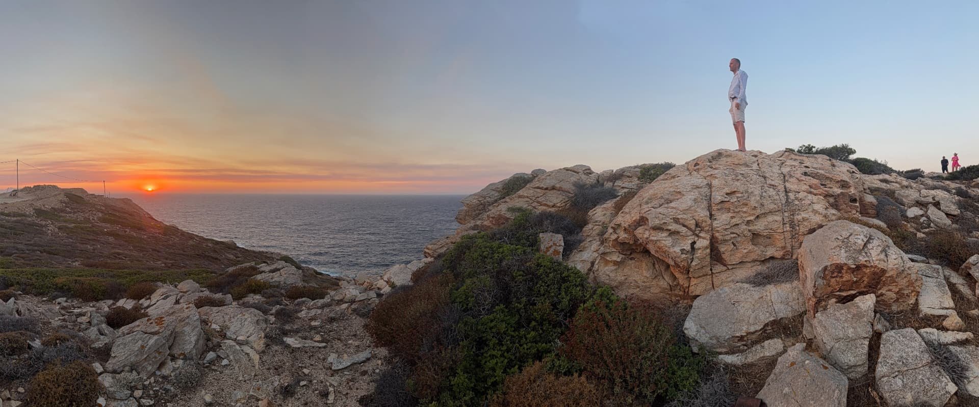 Wide panorama of the Akri House site at sunset, with Øystein Tynning standing on a large rock above the sea.
