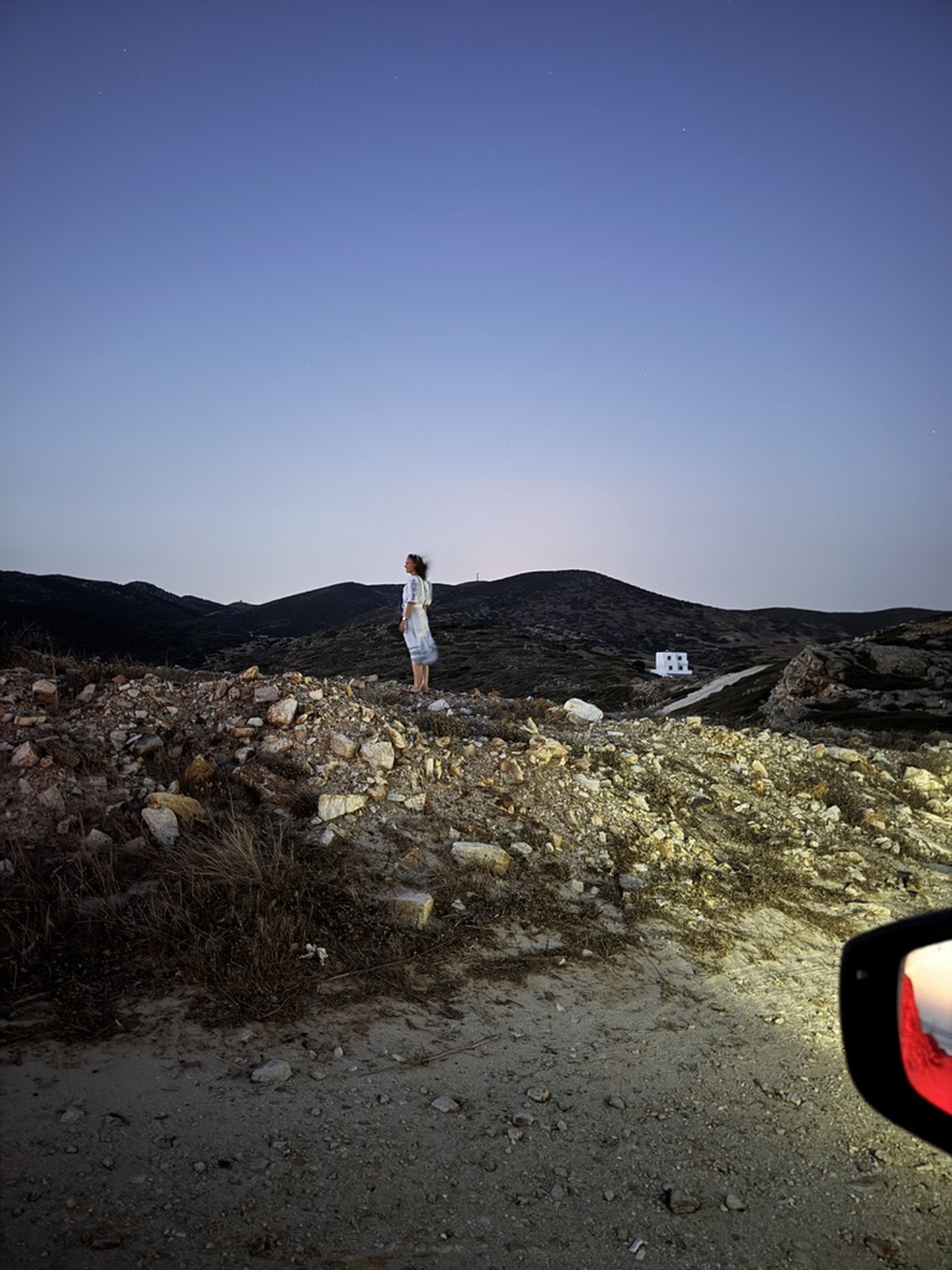 A person standing on the rocky Akri House site at dusk, with low hills in the distance and a car side-mirror in the foreground.
