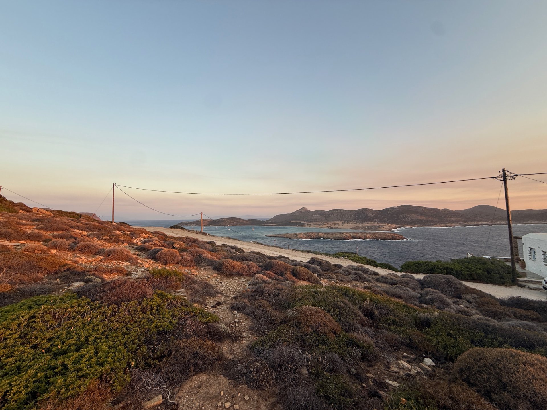 Low vegetation and exposed rock on the Akri House site, looking across the water towards the low hills beyond the bay.